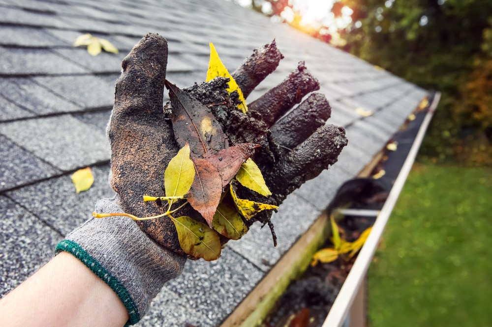 A gloved hand clears debris from a gutter along a roof