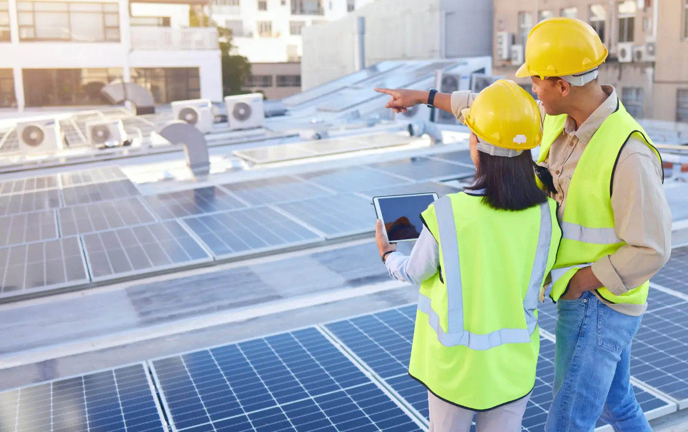 Two engineers wearing safety helmets and reflective vests inspect solar panels on a roof, one points and the other holds a tablet.